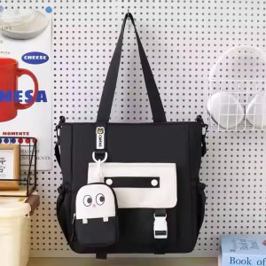 Black canvas tote shoulder bag with a white front pocket and a cartoon-face charm, hanging on a pegboard — Electromart Uganda
