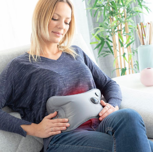 Grey rechargeable electric hot water bottle warming a woman's stomach on a sofa — Electromart Uganda